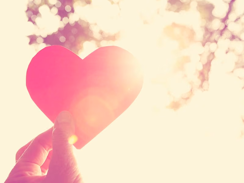 Hand holding a red paper heart with bright sunlight and bokeh in the background.