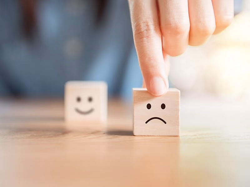 Finger pressing a wooden block with a sad face, with a happy face block blurred in the background.