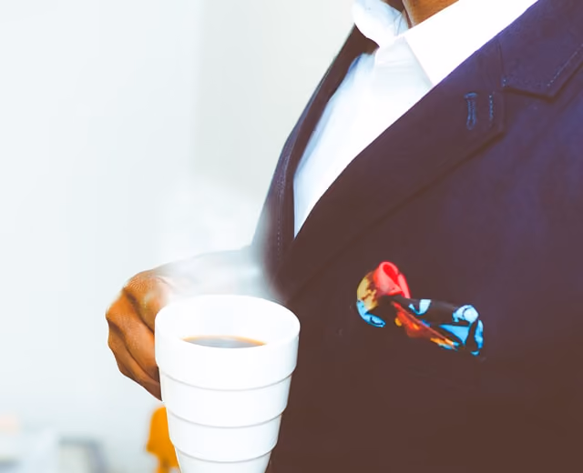 Person in a dark suit holding a steaming white cup, with a colorful pocket square visible.