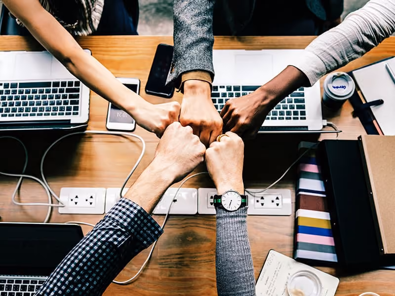 Five people joining fists in a teamwork gesture over a cluttered office desk with laptops and notebooks.