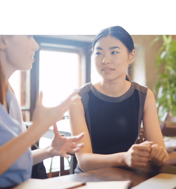 Two women having a focused conversation at a wooden table, one listening attentively to the other.