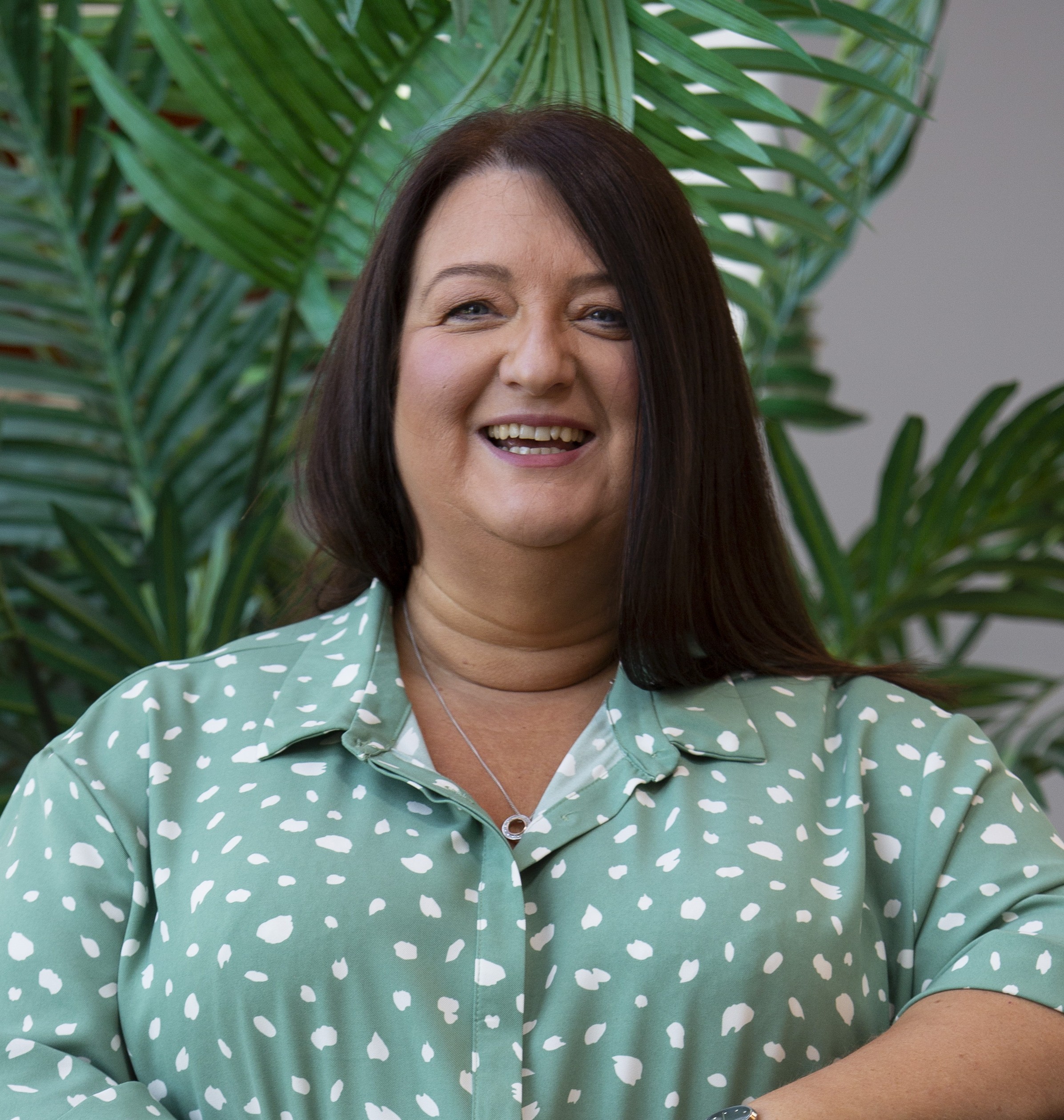 Smiling woman with long dark hair wearing a green and white patterned shirt in front of leafy plants.