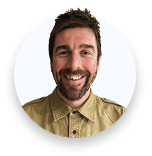 Man with short brown hair and beard smiling, wearing a tan buttoned shirt against white background.