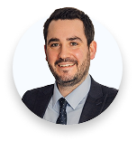 Smiling man with short dark hair, beard, wearing dark suit, light shirt, and dark tie against white background.