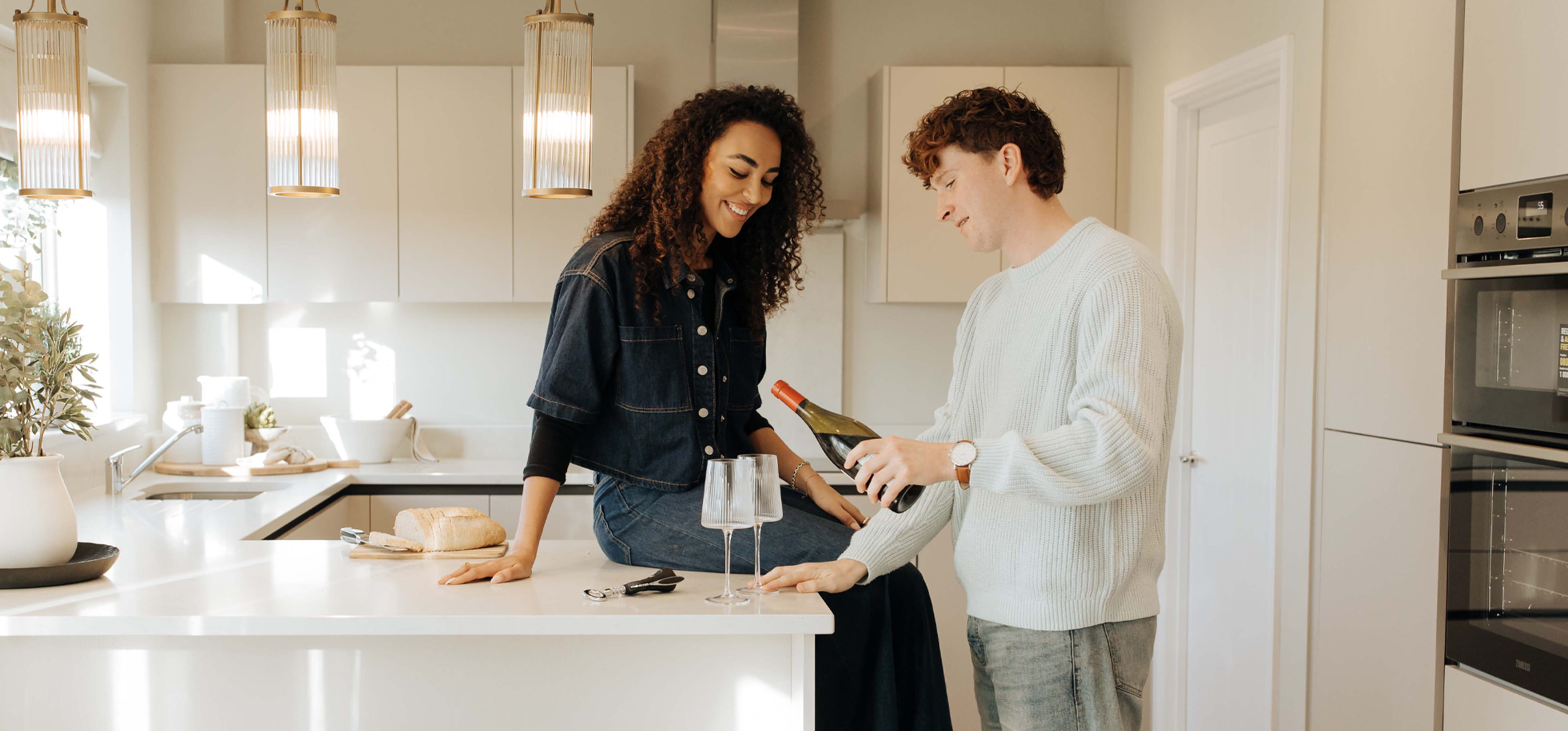 A woman with curly hair sitting on a kitchen counter smiling at a man pouring wine into two empty glasses in a modern, bright kitchen.