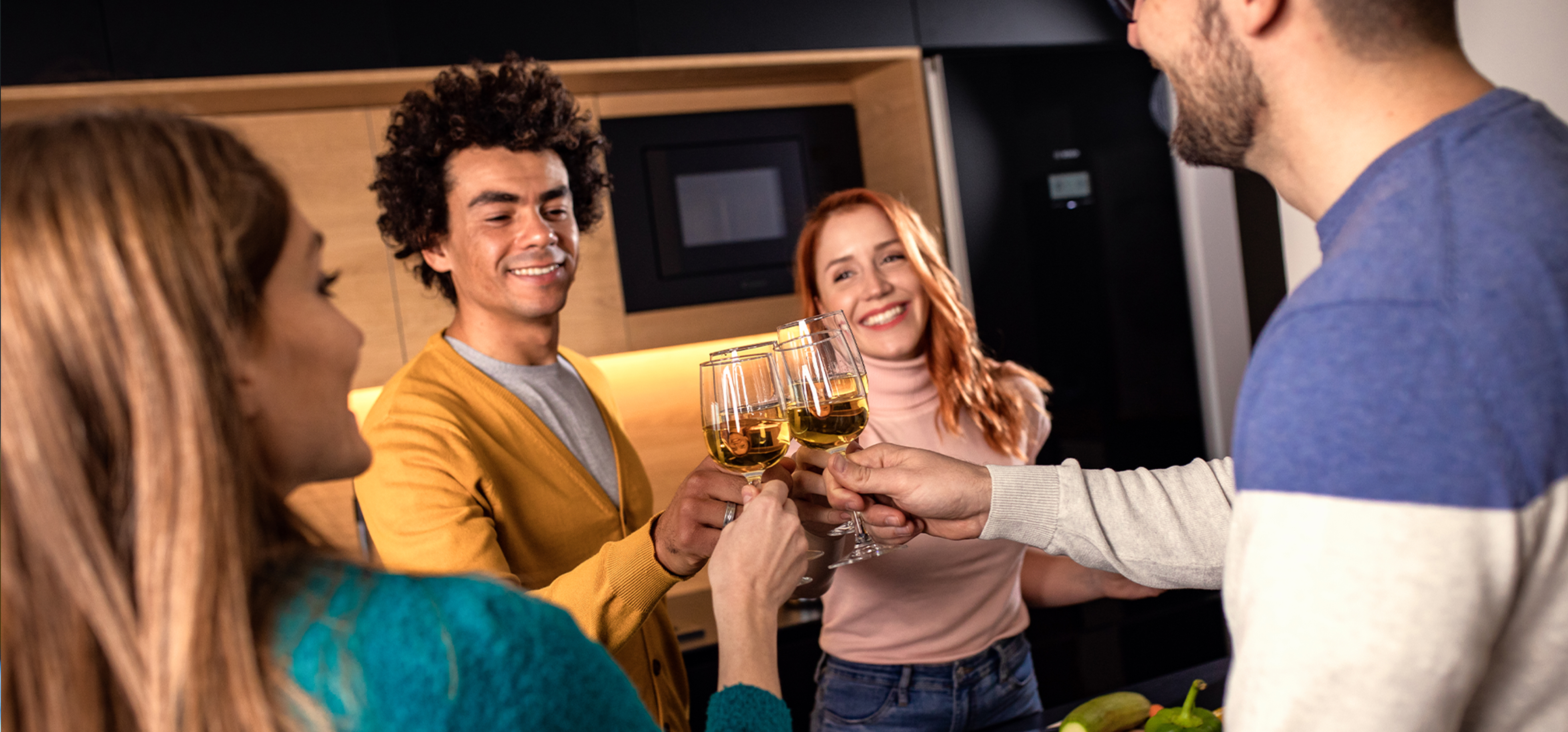 Four friends smiling and clinking glasses filled with white wine in a kitchen setting.