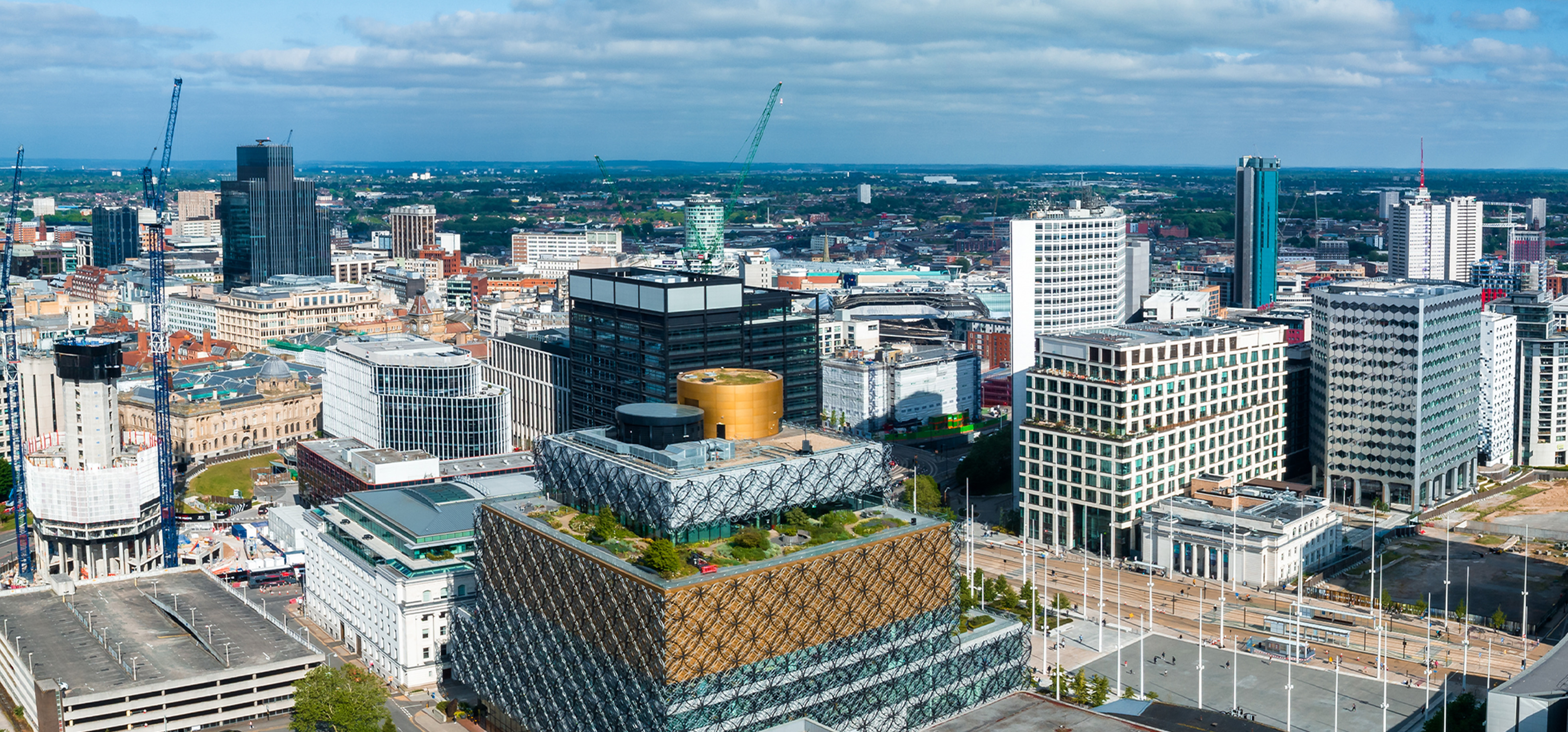 Aerial view of a modern cityscape with a prominent building featuring a distinctive patterned facade and rooftop garden in the foreground, surrounded by various office buildings and cranes under a partly cloudy sky.