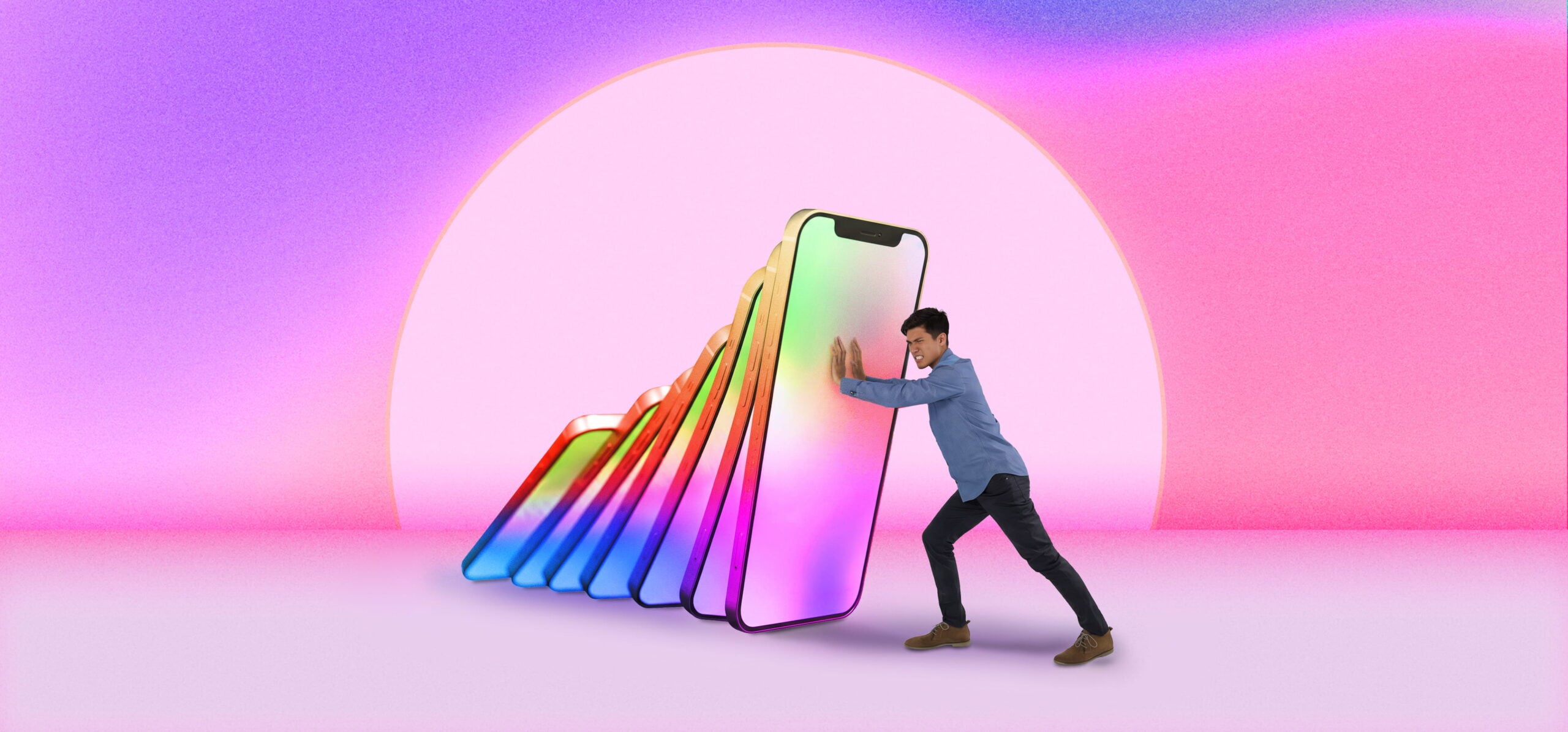 A man in a blue shirt and dark pants is pushing against a large upright smartphone with a colorful screen, preventing a row of similarly large smartphones from toppling like dominoes against a pink and purple gradient background with a large circular light behind.