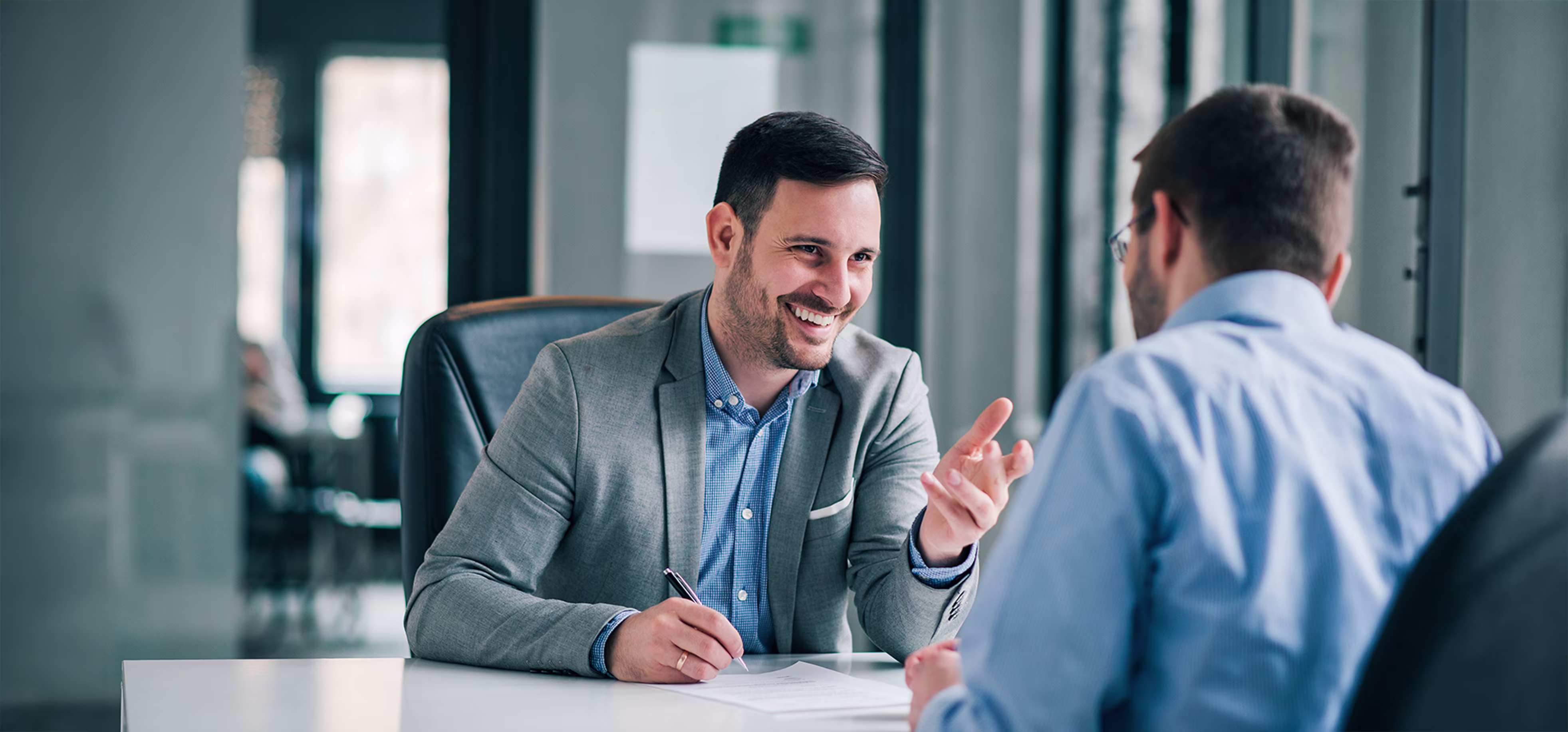 Two men in a professional office setting engaged in a friendly conversation across a white table, one man in a grey blazer smiling and gesturing while holding a pen, the other man seen from behind in a light blue shirt.