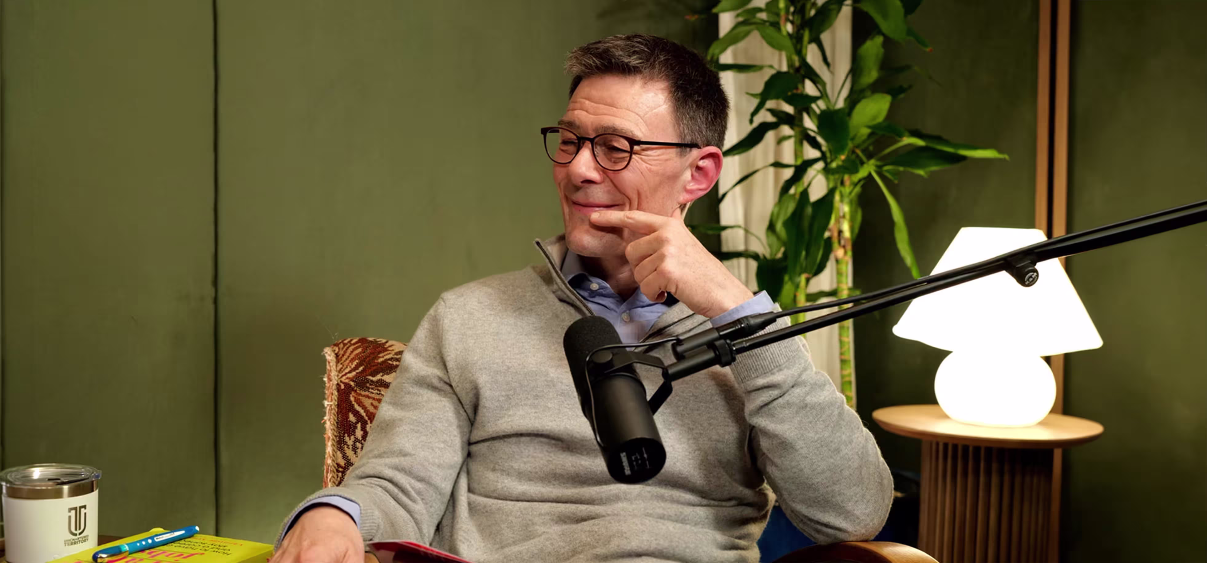Man wearing glasses and a grey sweater sitting in a chair with a microphone in front of him, pointing and smiling, against a green wall background with a plant and a lamp on a table.
