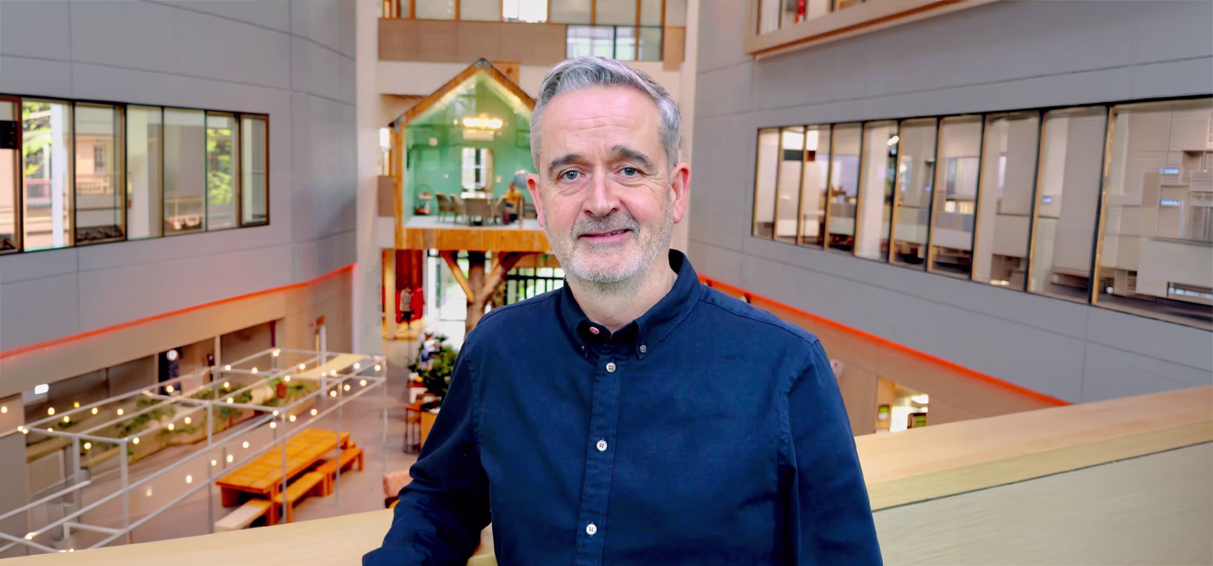 A middle-aged man with grey hair and beard wearing a dark blue buttoned shirt, standing indoors in a modern office building with a spacious atrium, glass walls, wooden benches, and a small house-like structure in the background.