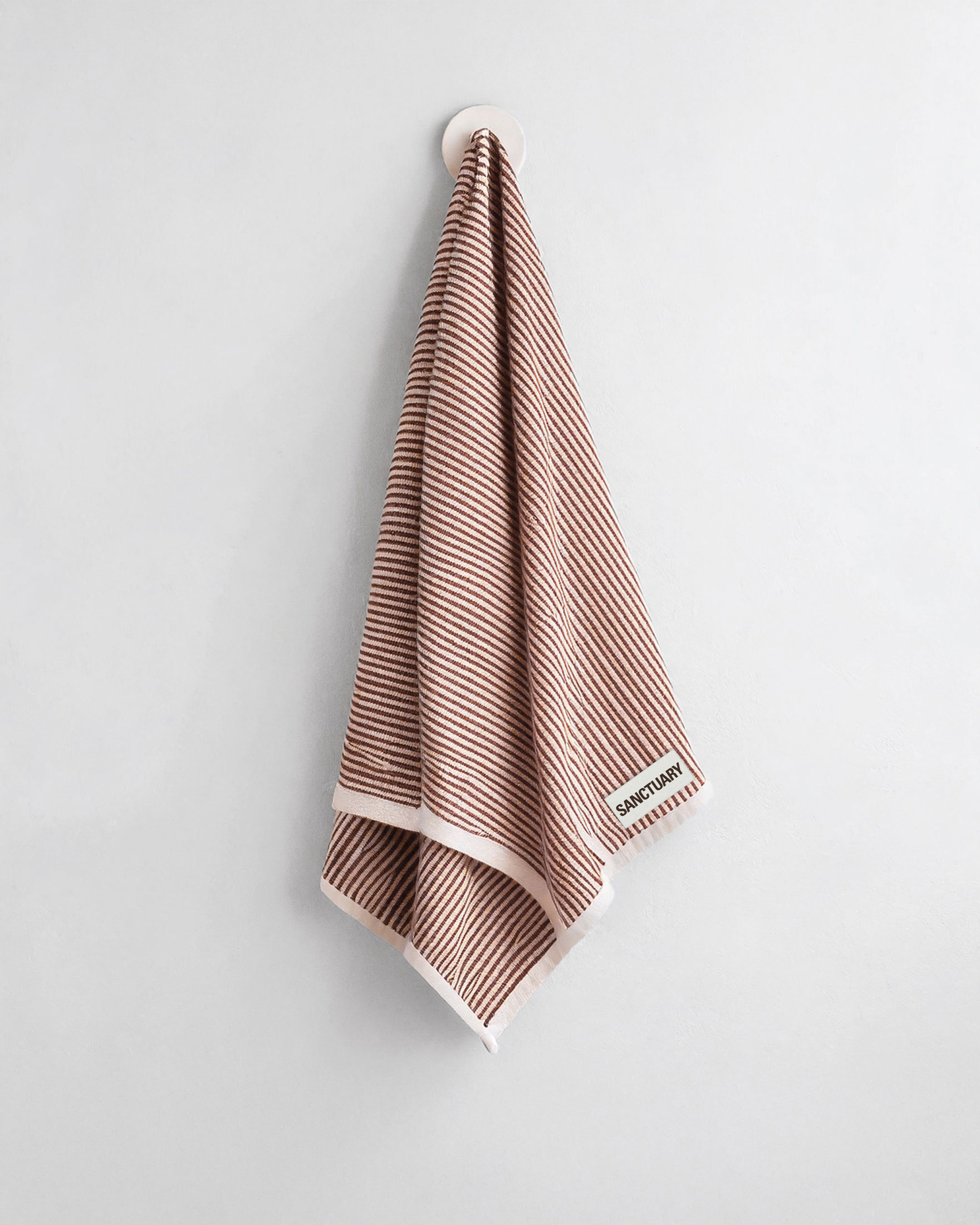 Brown towel with a small Sanctuary brand label on a pale wall
