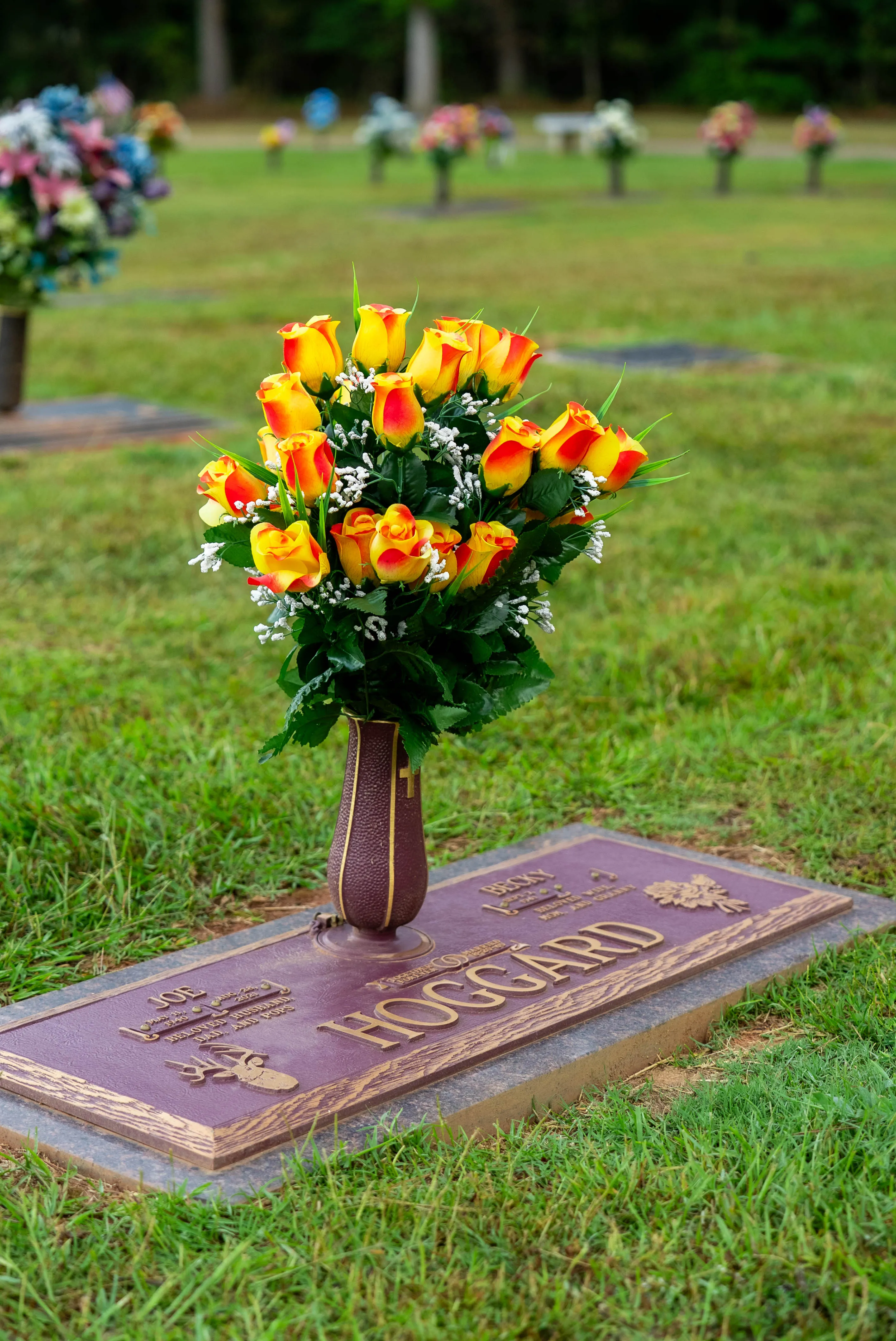 Bronze flat grave marker with a vase of orange and yellow roses in a cemetery lawn