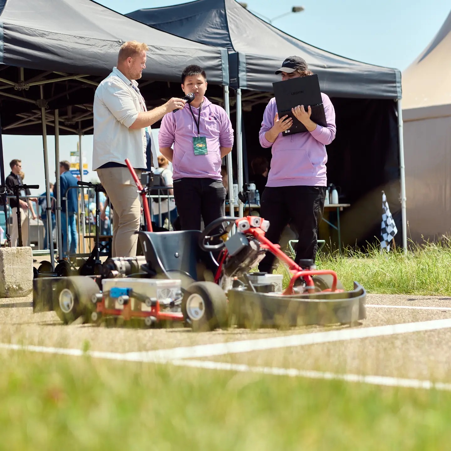 Interview met deelnemers naast een zelfrijdende kart, technische apparatuur en tenten buiten op een zonnige dag.