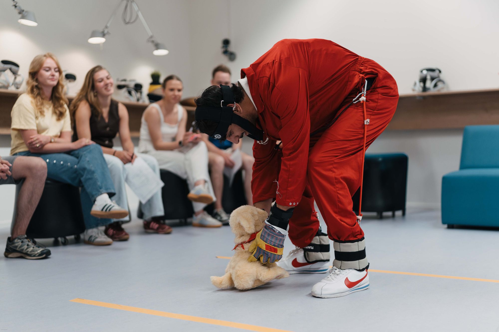 Persoon met verzwaringspak buigt naar een kleine labradorpup in een kamer met zittend publiek.