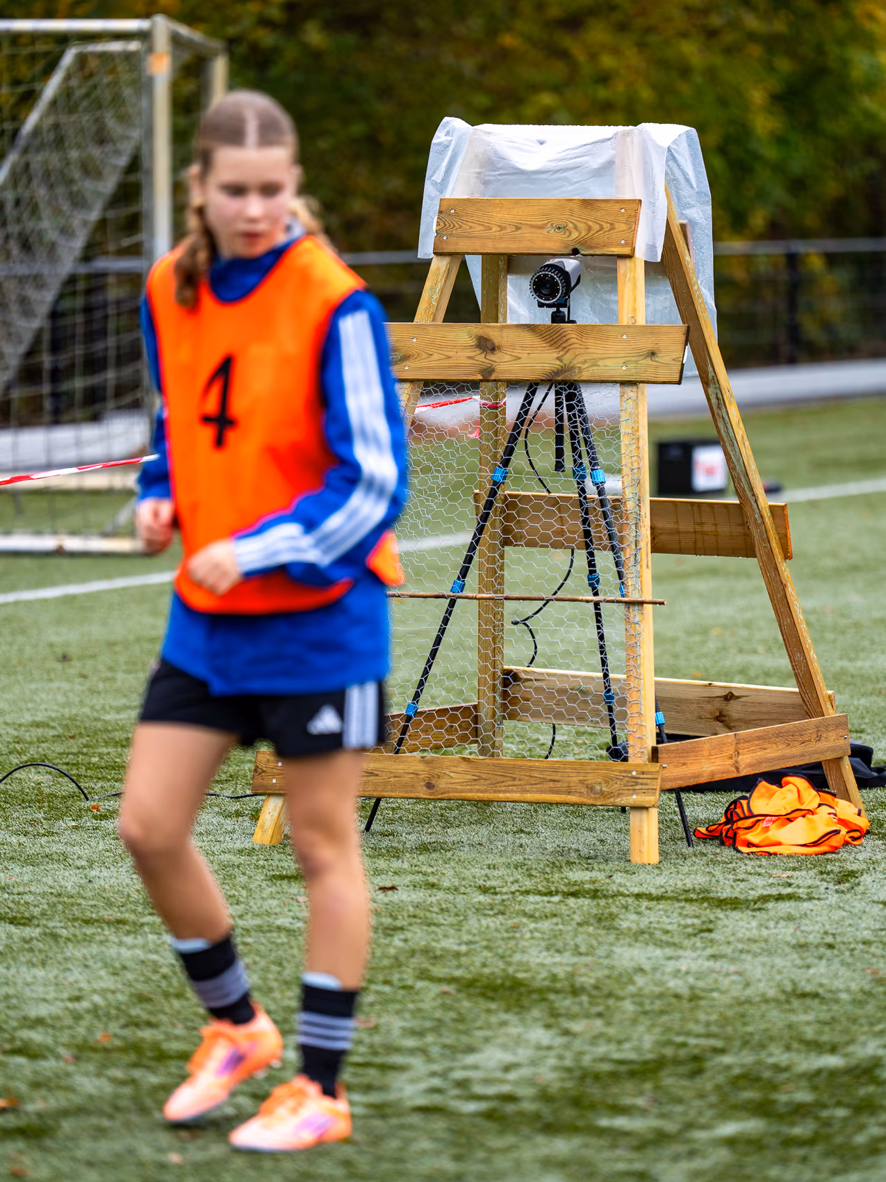 Meisjesspeler met oranje hesje op grasveld voor een houten statief met camera.