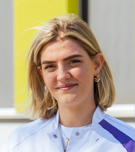 Jonge vrouw met blond haar en oorbellen draagt een witte jas met paarse accenten en lacht.