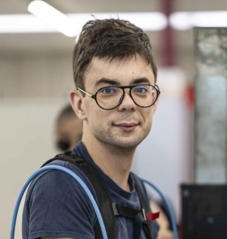 Jonge man met korte bruine haren en bril, draagt een blauw shirt en een rugzak.
