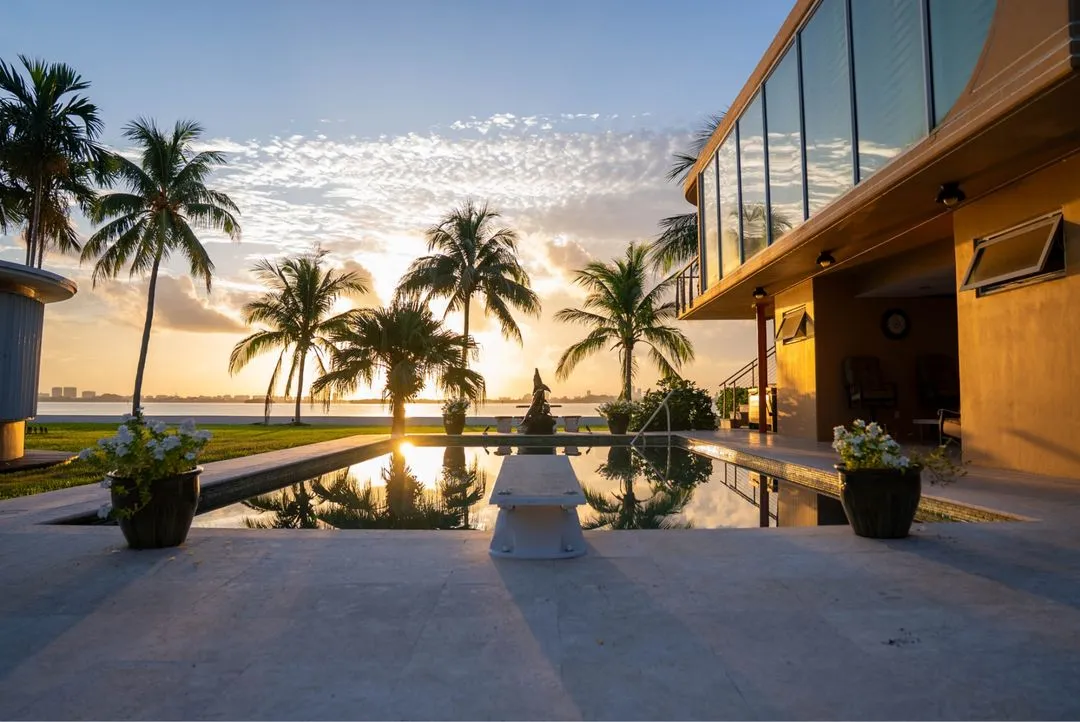 Palm trees silhouetted at sunset beside a backyard pool and diving board at a waterfront home