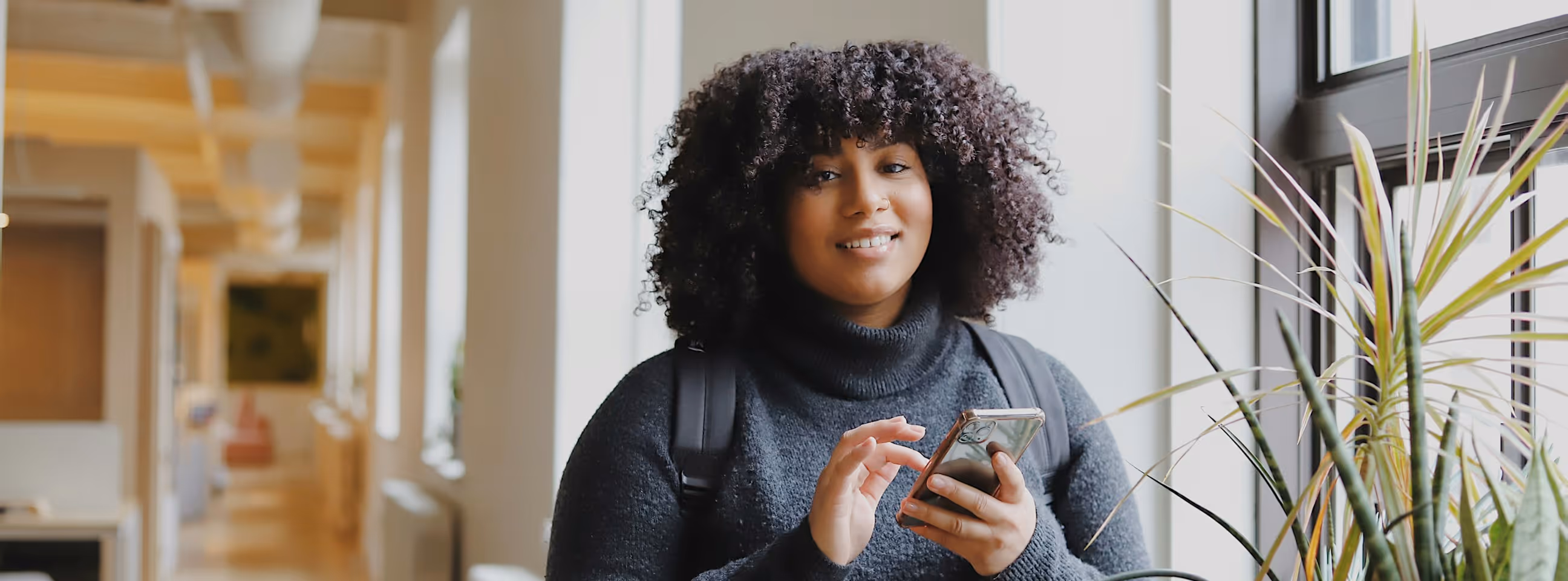 Jeune femme souriante avec sac à dos utilisant un smartphone près d'une fenêtre avec des plantes.