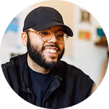 Homme souriant avec une barbe, portant une casquette noire et des lunettes assis à l'intérieur.