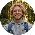 Jeune homme souriant avec un sweat à capuche gris et un sac à dos en forêt.