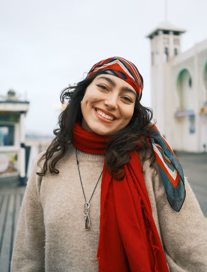 Jeune femme souriante portant un foulard coloré et une écharpe rouge sur un pull beige en extérieur.