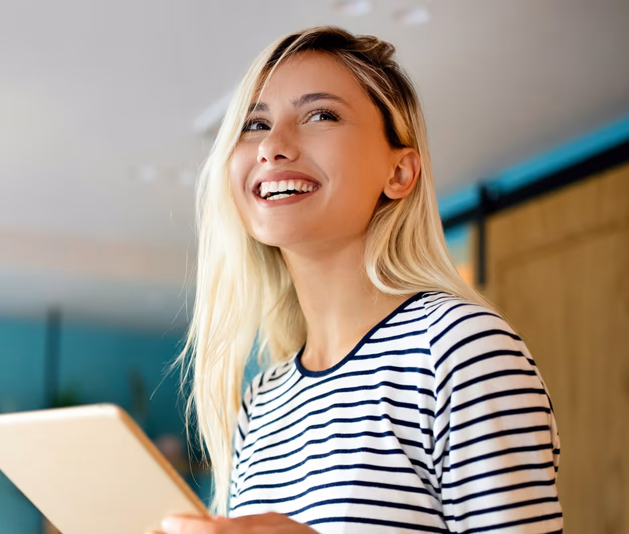 Jeune femme souriante aux cheveux blonds tenant une tablette avec un pull rayé blanc et noir.