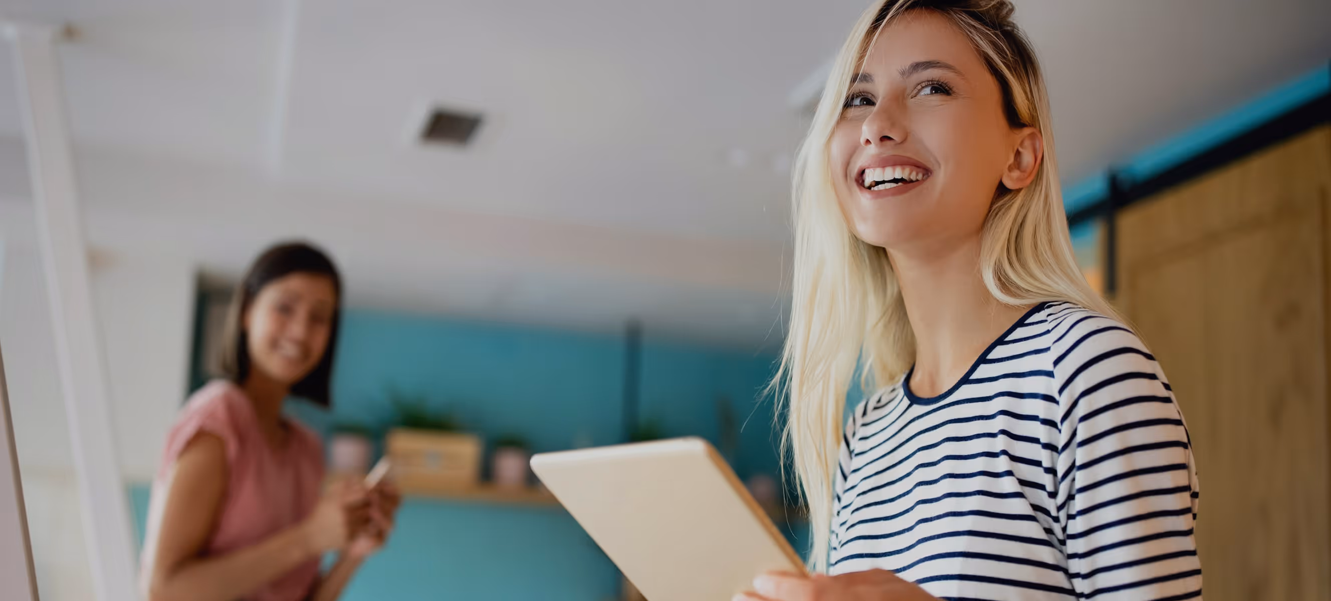 Jeune femme blonde souriante tenant une tablette, avec une autre femme floue en arrière-plan.