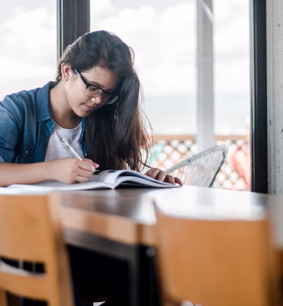 Une femme avec des lunettes écrit dans un cahier tout en étant assise à une table en bois.