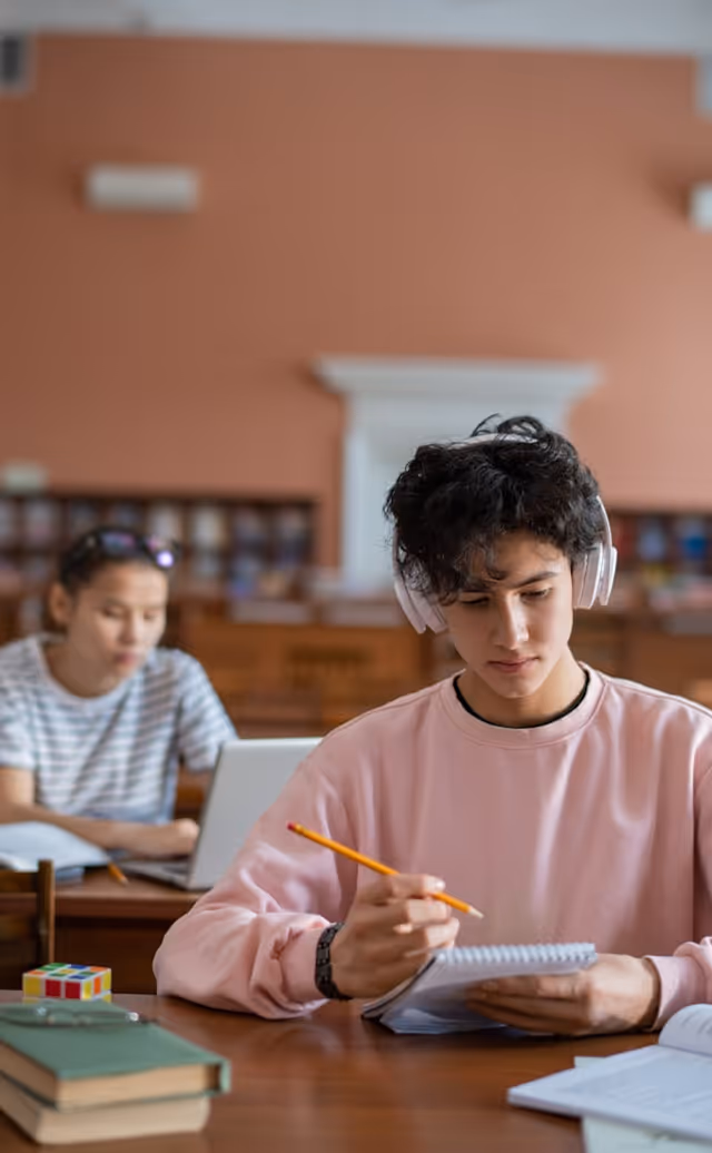 Un étudiant portant un casque écrit dans un cahier avec un autre étudiant et un ordinateur en arrière-plan.