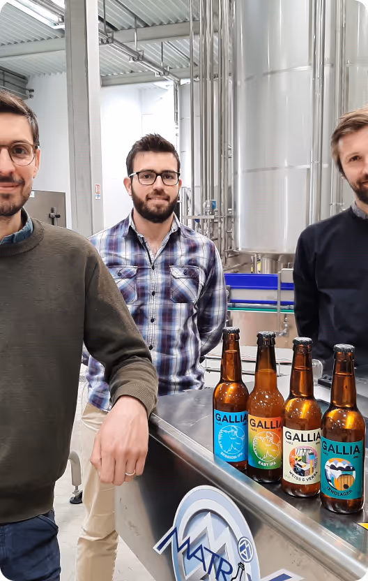 Trois hommes souriants debout près de bouteilles de bière Gallia sur une table en inox dans une brasserie.