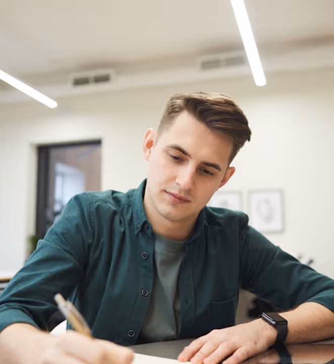 Jeune homme concentré écrivant dans un carnet à l'intérieur d'un bureau moderne.
