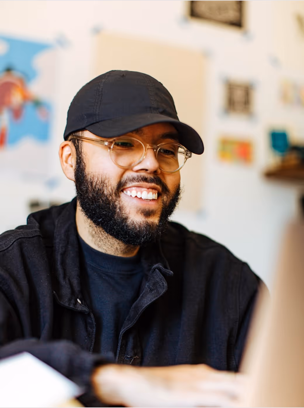 Homme souriant avec une casquette noire et des lunettes, travaillant sur un ordinateur portable.