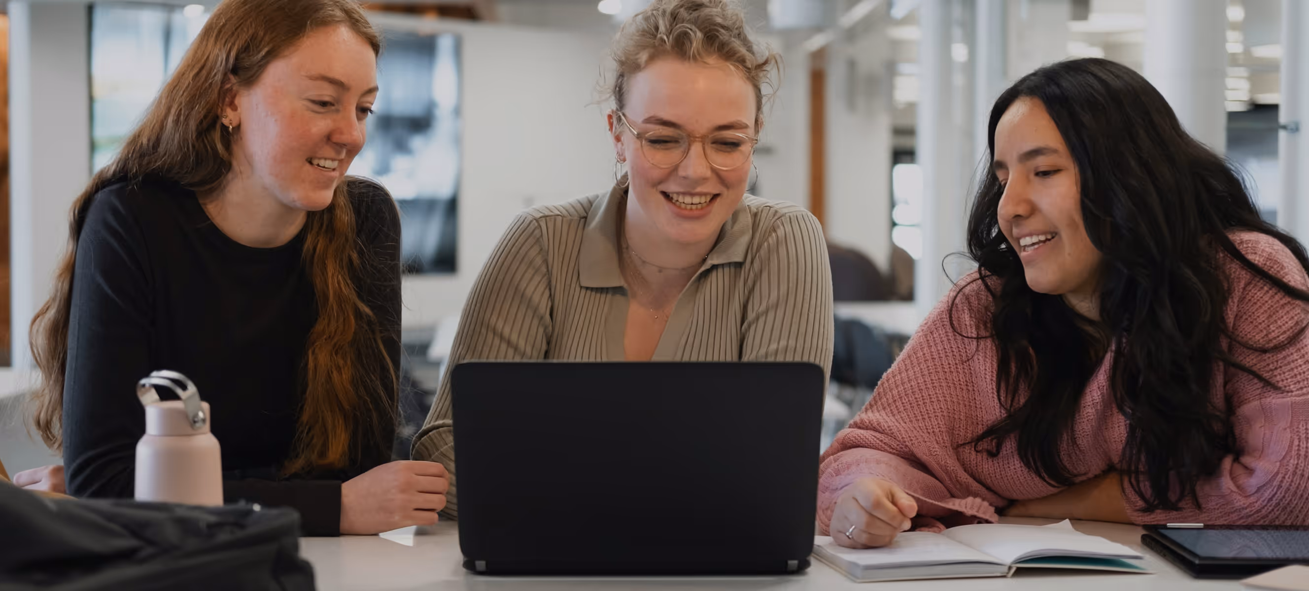 Trois jeunes femmes souriantes regardant un ordinateur portable ensemble dans un cadre de travail moderne.
