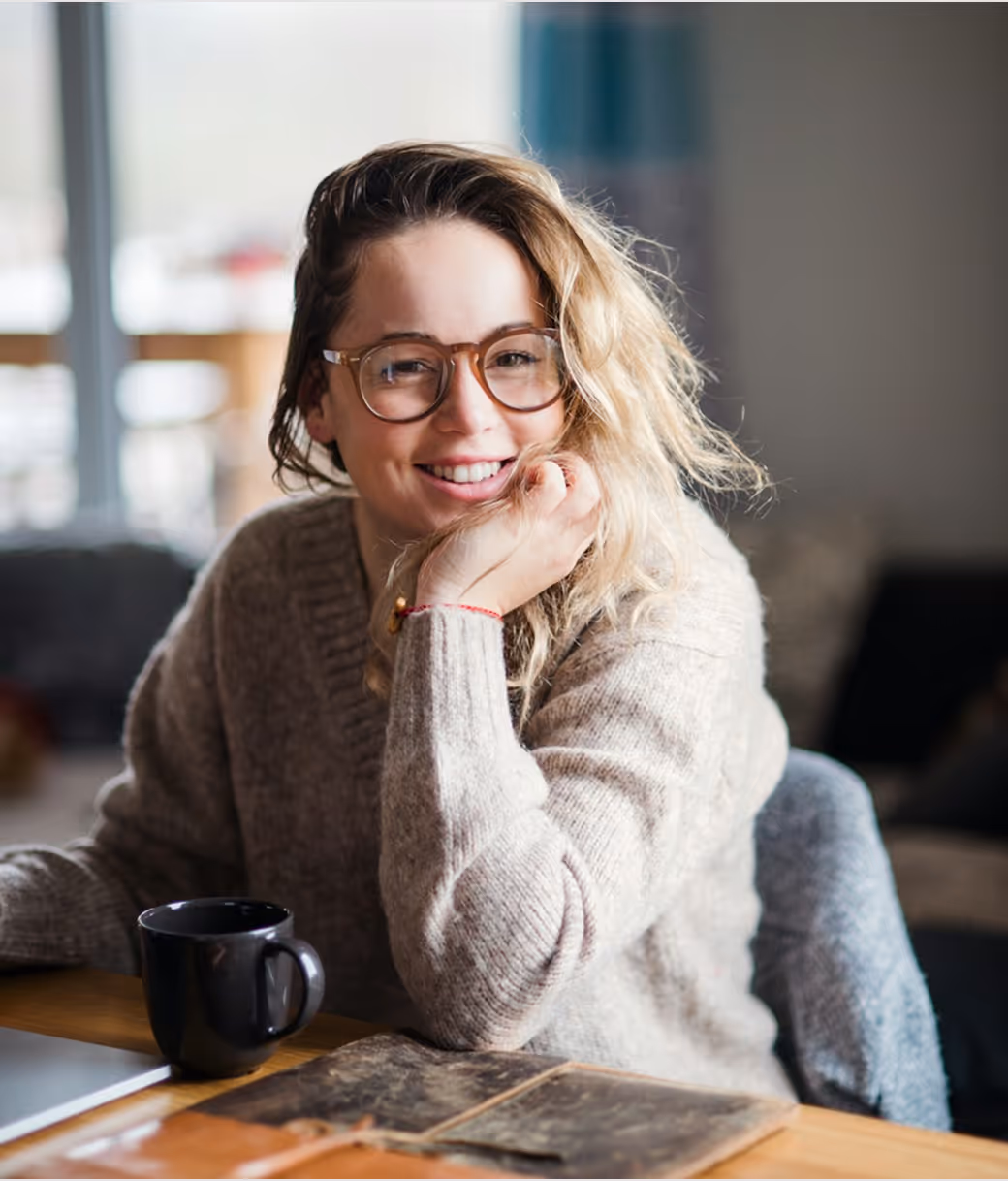Femme souriante avec lunettes, pull beige, assise à une table avec une tasse noire et un carnet ouvert.