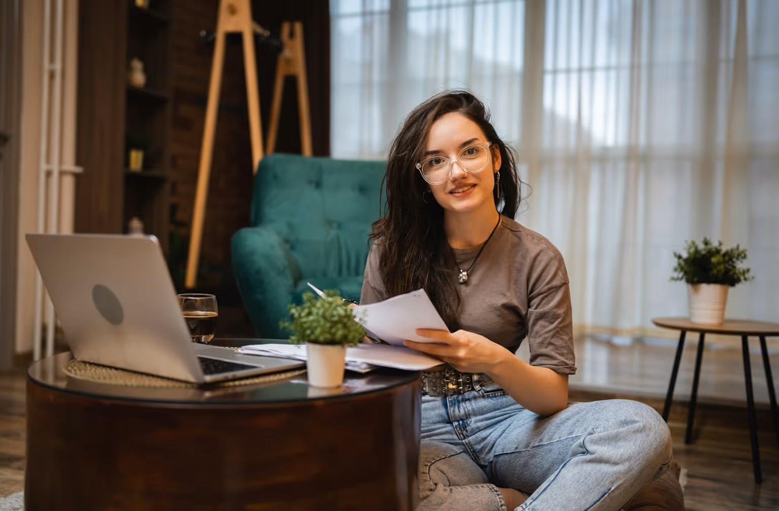 Jeune femme assise au sol avec des papiers, ordinateur portable et plantes dans un salon.