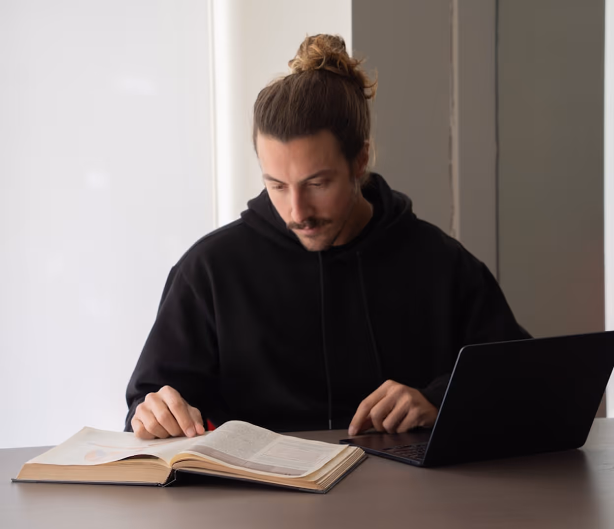 Homme lisant un livre ouvert à côté d'un ordinateur portable sur une table.