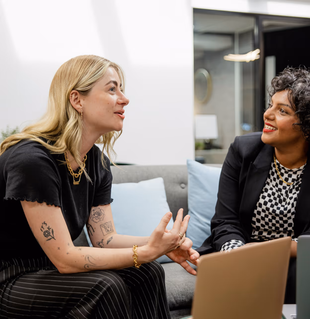 Deux femmes parlant ensemble assises sur un canapé gris dans un bureau moderne.