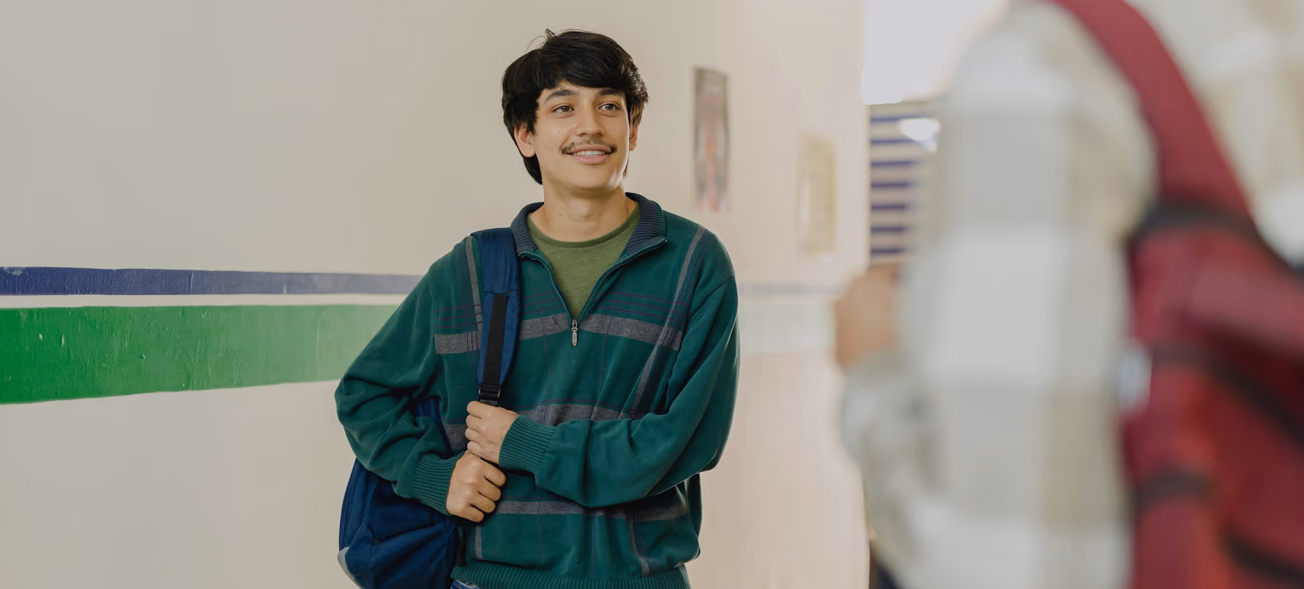 Un jeune homme souriant avec un sac à dos bleu debout dans un couloir lumineux.