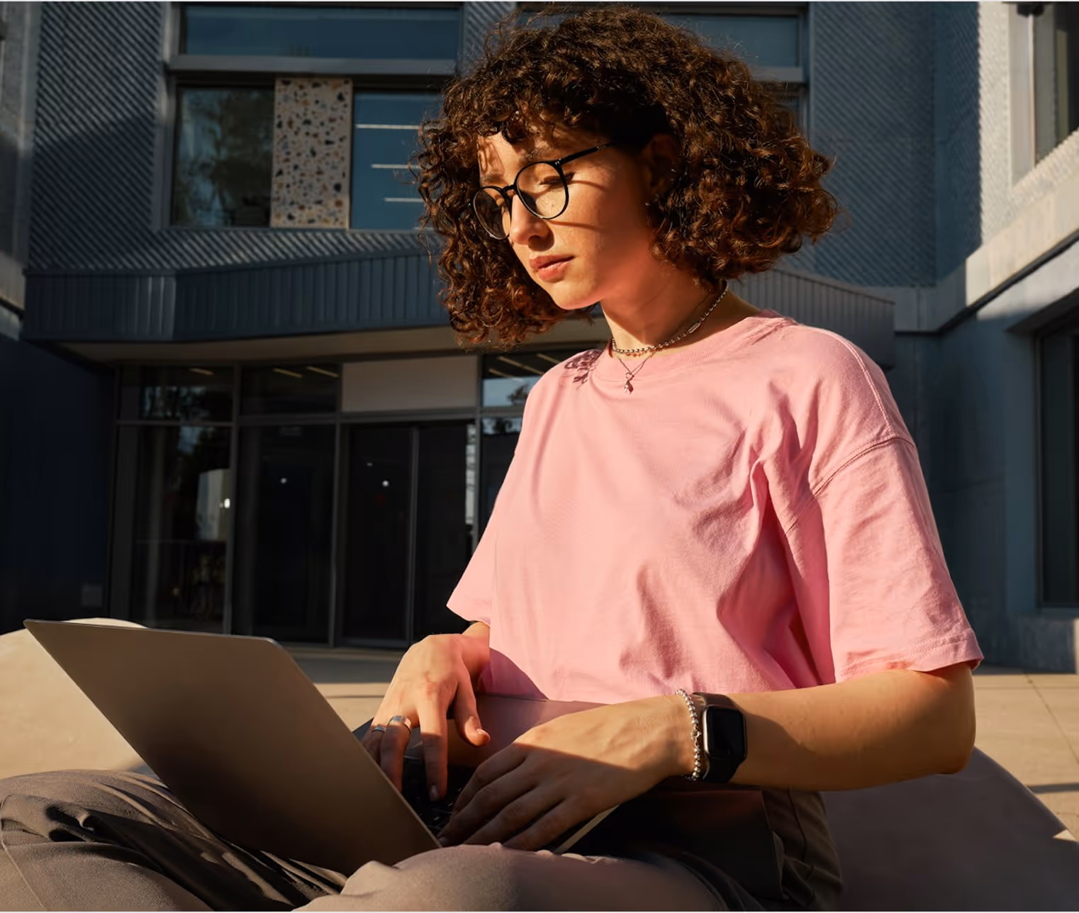 Jeune femme avec lunettes et t-shirt rose travaillant sur un ordinateur portable en extérieur.
