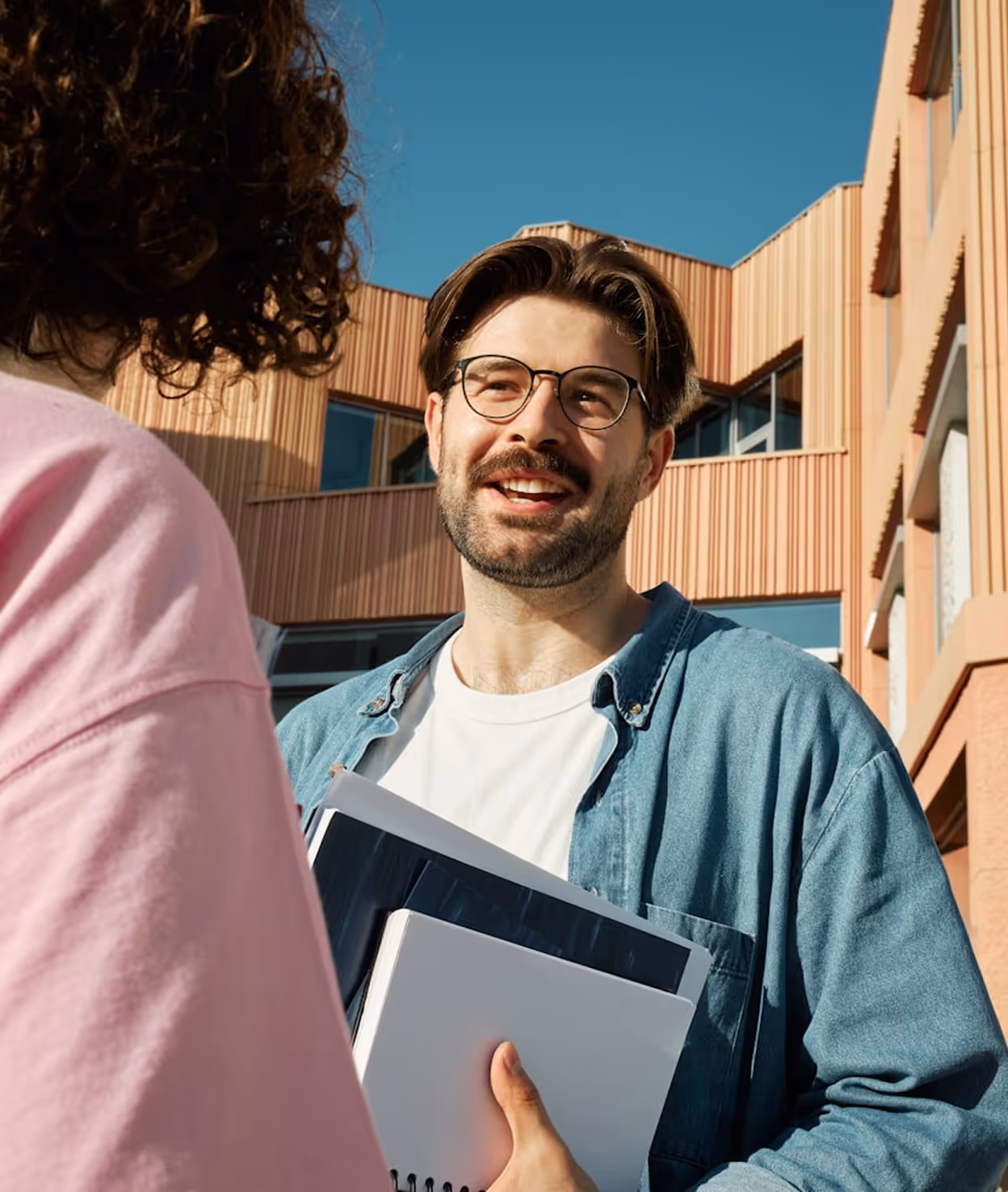 Un homme souriant avec des lunettes parle à une personne, tenant des dossiers devant un bâtiment.