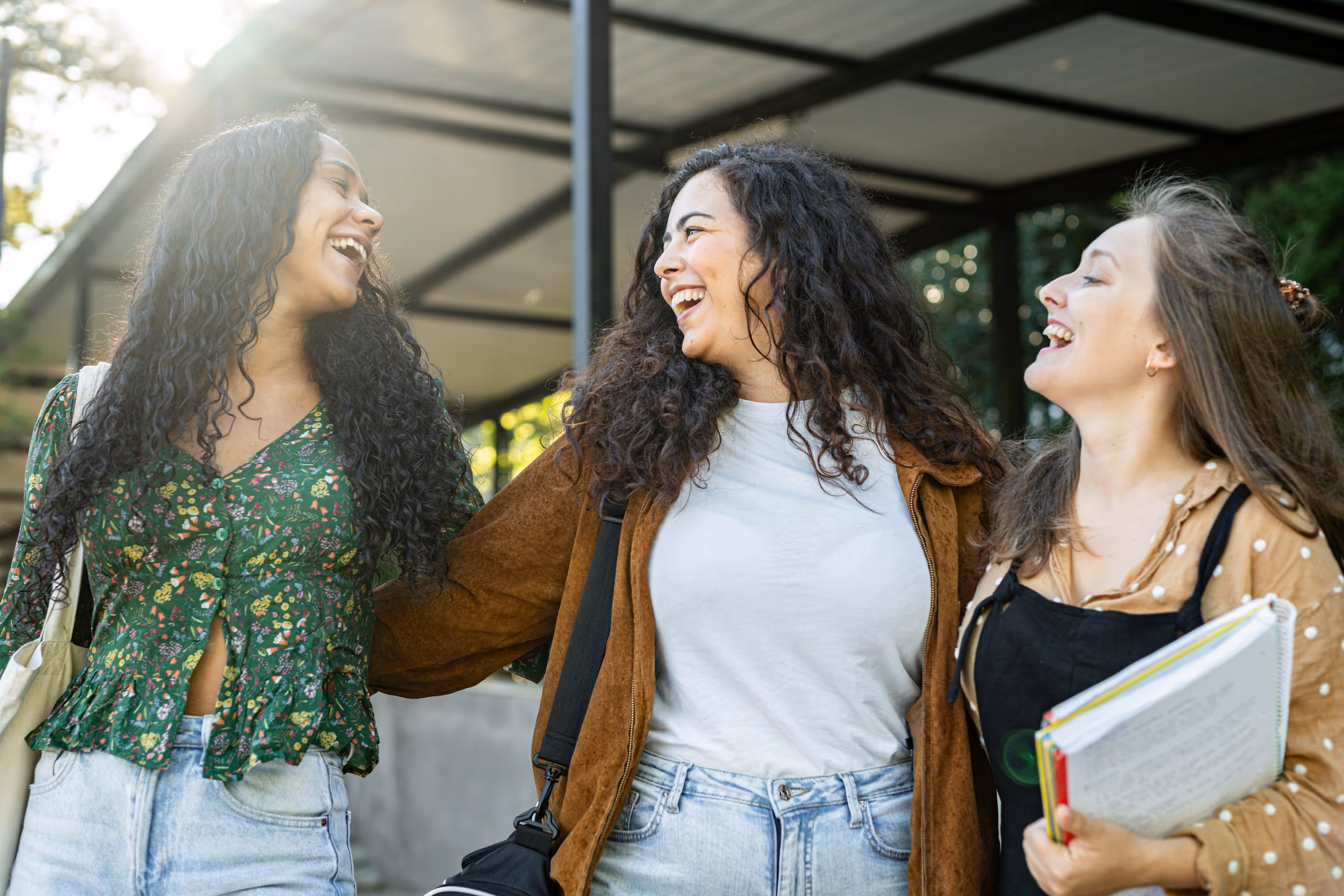 Trois jeunes femmes souriantes marchant ensemble dehors en tenant des livres et sacs.