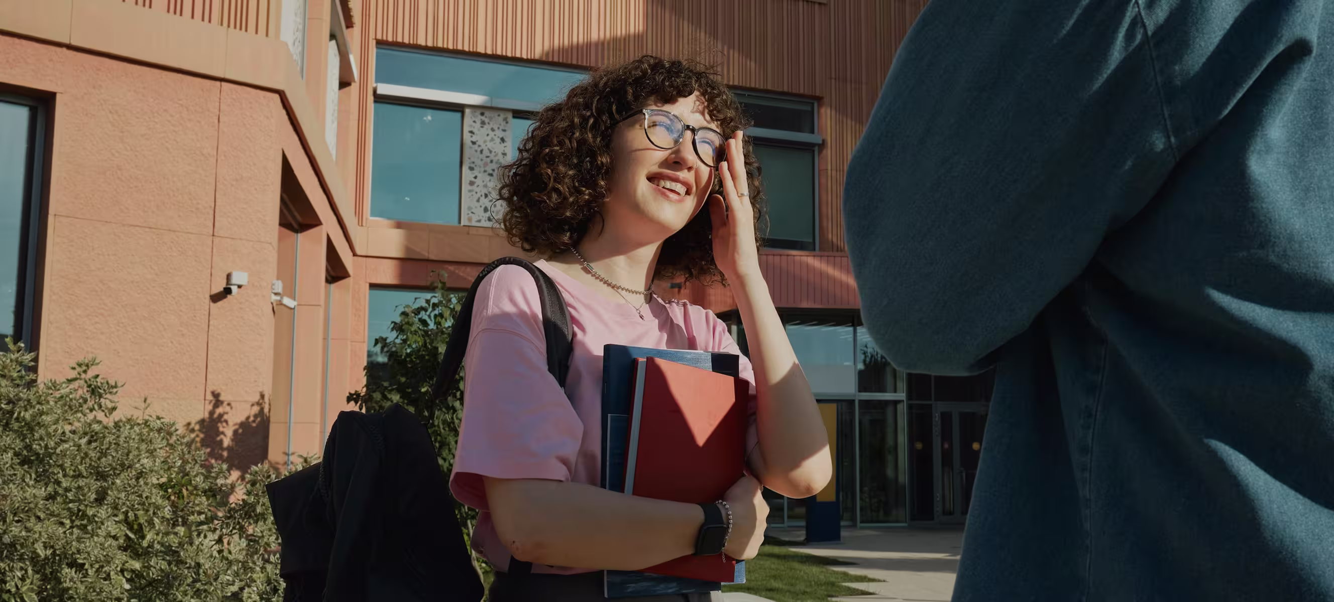 Jeune étudiante souriante portant un sac à dos et tenant des dossiers devant un bâtiment universitaire.