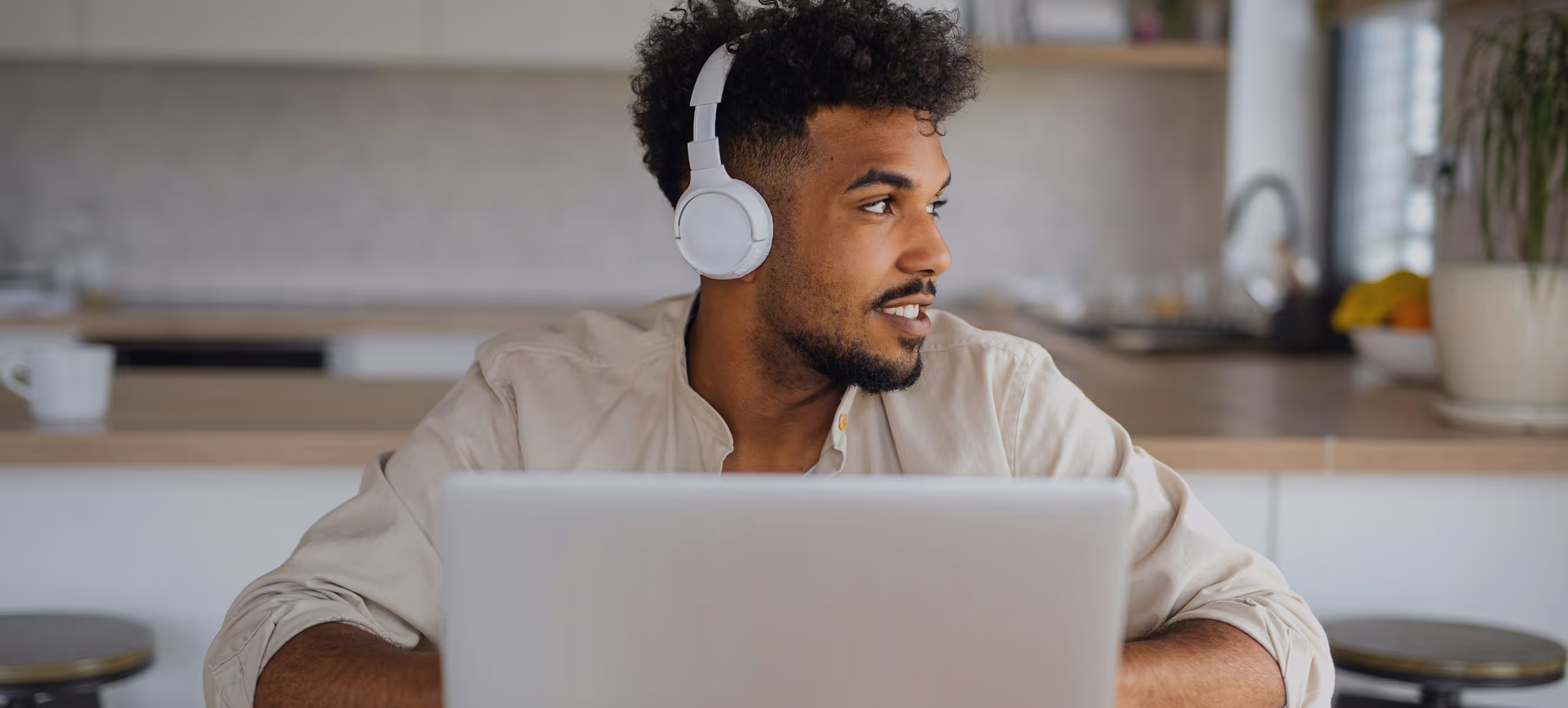Homme portant un casque blanc et souriant en travaillant sur un ordinateur portable dans une cuisine.