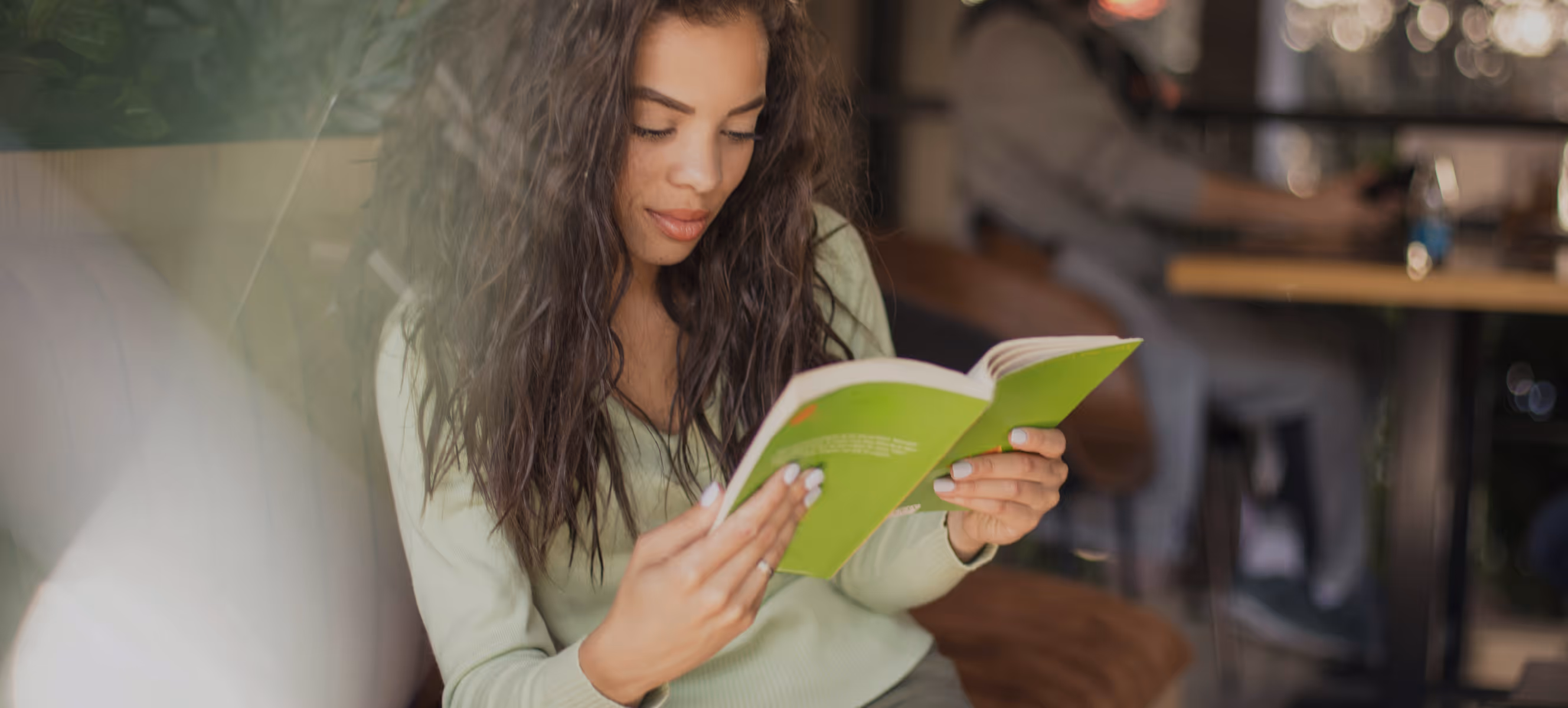 Jeune femme cheveux bouclés lisant un livre vert dans un café moderne.