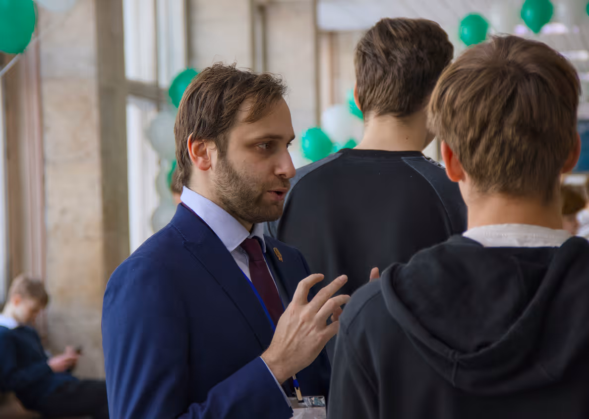 Un homme en costume bleu parle avec deux jeunes vêtus de noir lors d'un événement décoré de ballons verts.