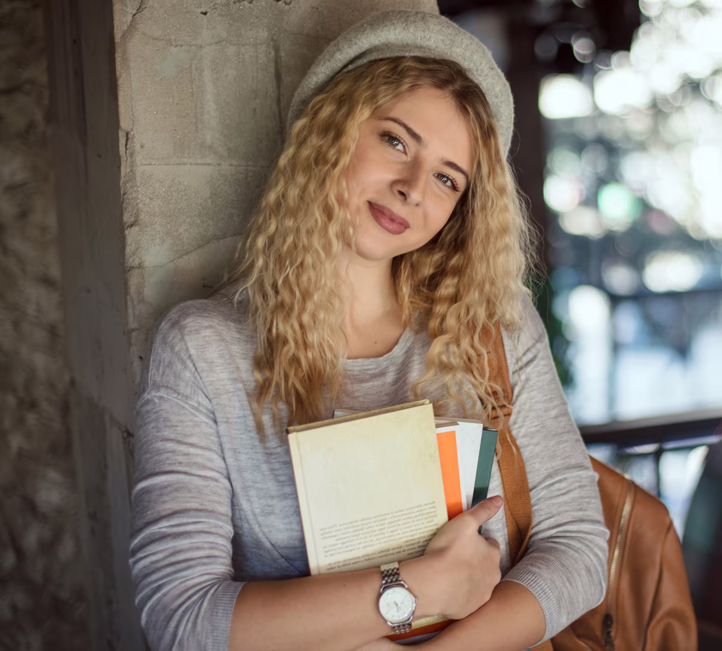 Jeune femme souriante aux cheveux bouclés tenant des livres contre un mur en béton.