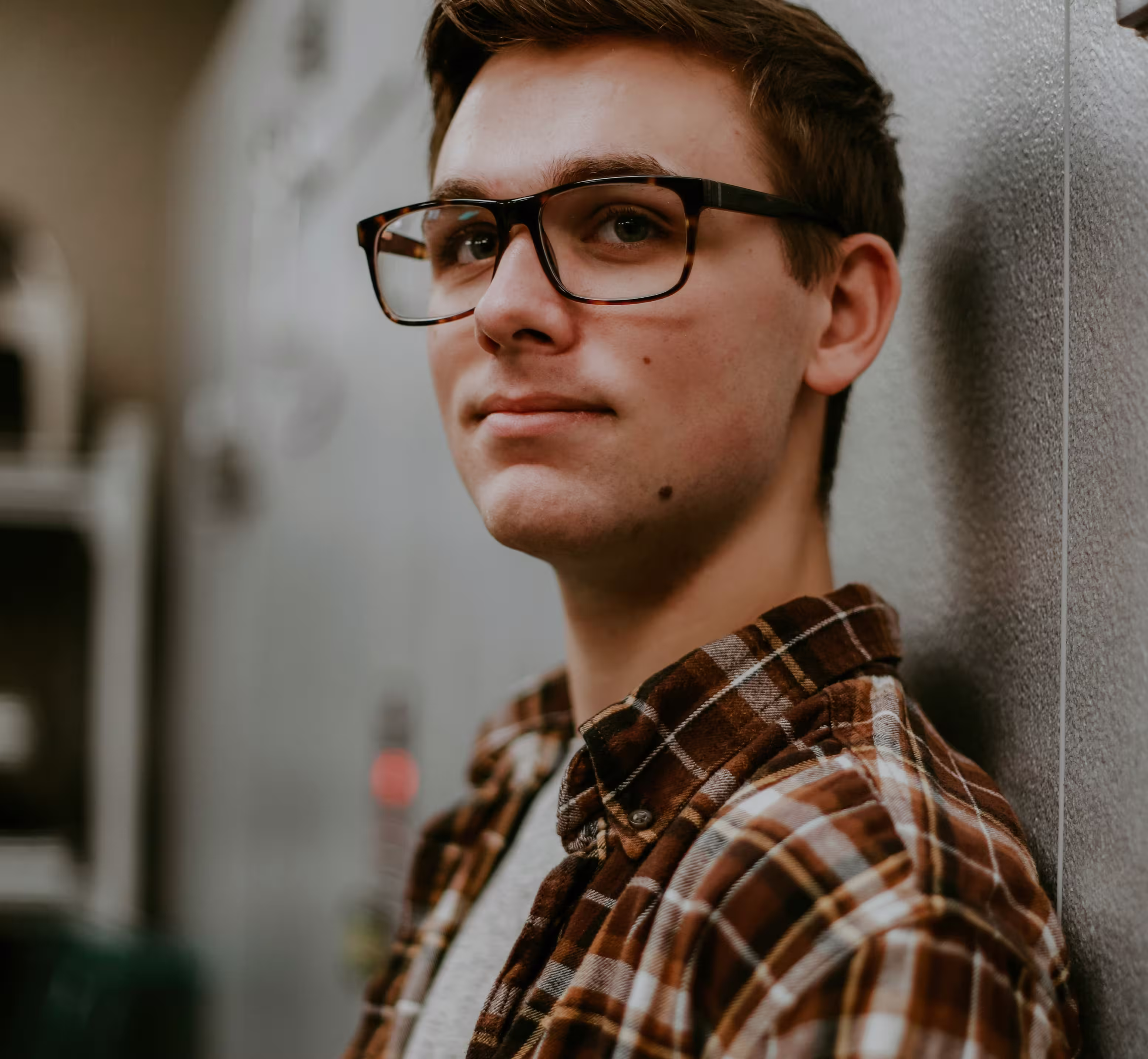 Jeune homme avec lunettes et chemise à carreaux marron, appuyé contre un mur gris.