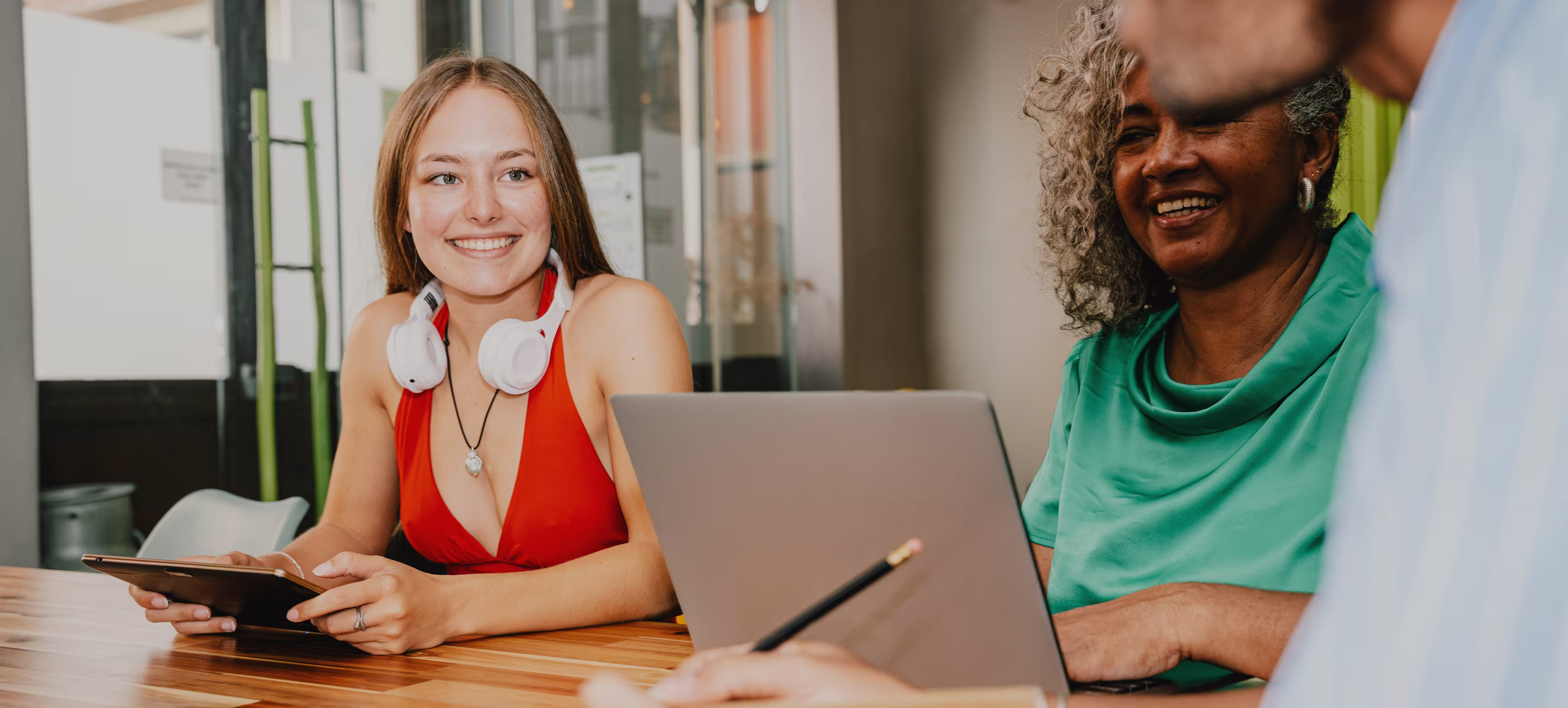 Trois personnes souriantes discutent en groupe avec ordinateur portable et tablette sur une table en bois.